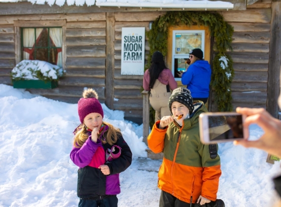 Two children have their picture taken while they eat maple syrup off of popsicle sticks in front of a wooden cabin in the winter.