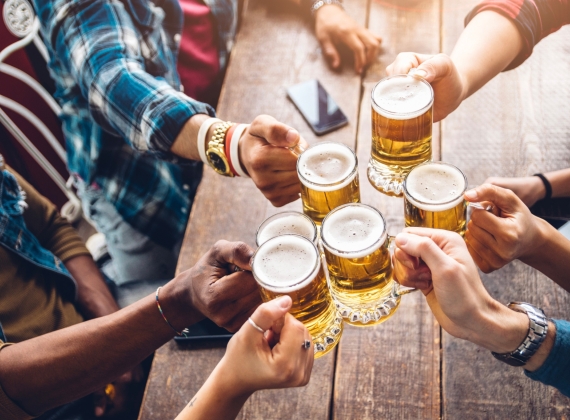 Overhead view of 6 people clinking beers on a wooden table with warm sunshine.