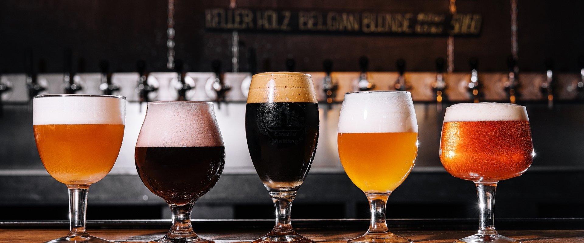 5 different beers in a range of styles of glasses on a wooden bar top.